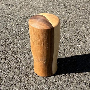 May include: A handcrafted wooden shift knob with a two-tone design. The top is split into two sections, one dark brown and the other light brown. The body is a light brown color. The knob is cylindrical with a slightly curved base.