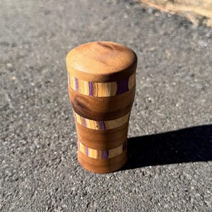 May include: A handcrafted wooden bottle stopper. The cylindrical stopper has a rounded top. It features alternating bands of dark and light wood, with thin purple stripes. The wood grain is visible, and the stopper is likely for wine bottles.