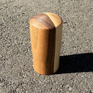 May include: A cylindrical wooden object with a rounded top. The top is divided into two sections, one dark brown and the other light brown. The body is a light brown colour. The object is standing on a grey surface.