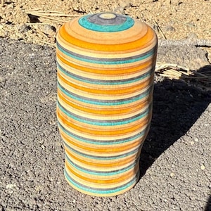 May include: A cylindrical wooden vase with a rounded top. The vase is made of layered wood in shades of orange, teal, gray, and natural wood tones. The vase is standing on a rough, textured surface.