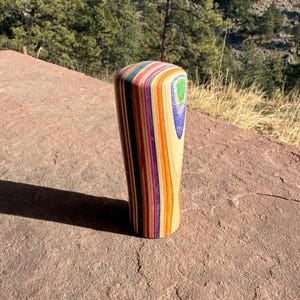 May include: A wooden, cylindrical object with a rounded top. The body is composed of colorful, layered wood strips in shades of red, orange, yellow, purple, and blue. A small, curved section on the side features green and blue accents. The object is set on a reddish-brown stone.