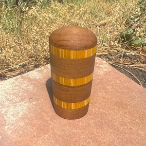 May include: A cylindrical wooden container with a rounded top, featuring alternating horizontal bands of dark brown wood and bright yellow wood with thin light-colored vertical stripes integrated into the yellow sections of the object.