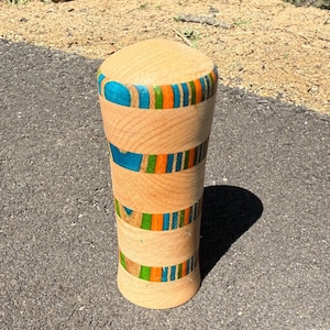Puede incluir: Un jarrón de madera con un acabado marrón claro. El jarrón presenta bandas de acentos azules, naranjas y verdes. El jarrón es cilíndrico con una parte superior redondeada y está sobre una superficie gris.
