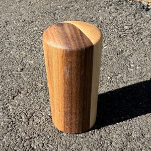 May include: A cylindrical wooden object with a flat top and a slightly tapered body. The wood has a natural grain and a two-tone colour scheme, with a darker brown section and a lighter brown section.