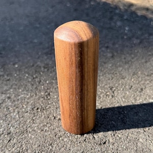 May include: A cylindrical wooden object with a rounded top. The wood grain is visible, and the color is a warm brown. The object is standing upright on a textured surface, casting a shadow.
