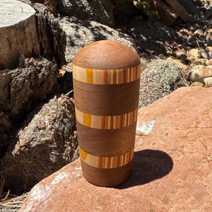 May include: A cylindrical wooden vessel with a rounded top, featuring alternating bands of dark brown wood and decorative segments of light wood inlaid with vertical stripes in shades of yellow, orange, and tan throughout the exterior surface.