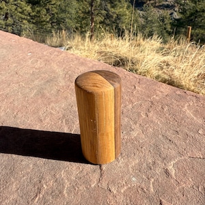 May include: A cylindrical wooden object, likely a bottle stopper, with a two-tone design. One half is light-colored wood, the other is a darker brown. The object is standing upright on a reddish-brown stone surface, with a natural outdoor background.