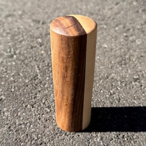 May include: A cylindrical wooden object, possibly a furniture leg or decorative item. It's made from two types of wood, one light and one dark, forming a contrasting vertical stripe. The top is rounded. The object casts a shadow on the ground.