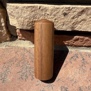 May include: A cylindrical wooden object with a rounded top. The wood grain is visible, and the color is a medium brown. The object is standing upright against a textured stone background.