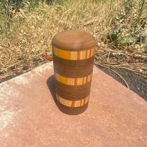 May include: A cylindrical wooden container with a rounded lid, featuring horizontal bands of dark brown wood alternating with decorative stripes of light tan, orange, and yellow wood grain patterns arranged in a repeating vertical design.