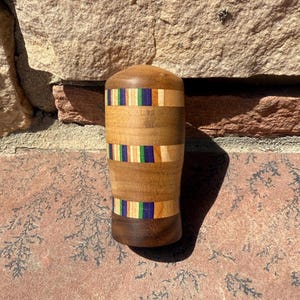 May include: A handcrafted wooden tumbler with a cylindrical shape. The tumbler is made of various wood tones, with bands of colorful inlaid wood featuring blue, green, and orange accents. The base and top are a darker wood.