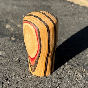 May include: A wooden shift knob with a layered design. The knob features alternating bands of light wood, black, and a thin red accent. The top is rounded, and the base is slightly wider. The object is sitting on a dark surface.