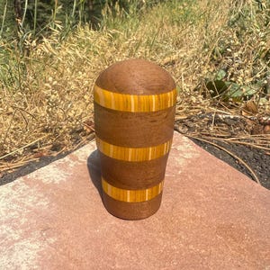 May include: A cylindrical wooden vessel with a rounded top, featuring alternating horizontal bands of dark brown wood and bright yellow wood with thin light-colored vertical stripes integrated into the yellow sections of the object.