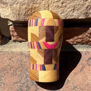 May include: A handcrafted wooden vase with a unique geometric design. The vase features alternating bands of light and dark wood, accented with vibrant pink, teal, and burgundy stripes. The vase is approximately 8 inches tall.