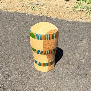 Puede incluir: Un jarrón de madera hecho a mano con una parte superior redondeada y un cuerpo cilíndrico. El jarrón presenta bandas de rayas turquesas, naranjas y verdes. El jarrón está hecho de madera de color claro.