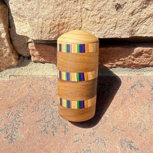 May include: A wooden container with a rounded top and a cylindrical body. The body features two bands of colorful, inlaid wood with blue, green, and orange accents. The container is made of light brown wood and sits on a reddish-brown surface.