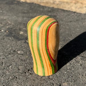 May include: A wooden shift knob with a rounded top and a curved base. The knob is made of layered wood, featuring alternating stripes of light brown, green, and red. The shift knob is designed for a vehicle.