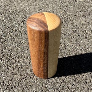 May include: A cylindrical wooden object, split vertically into two halves. One half is dark brown, the other is light tan. The top is rounded. The object rests on a textured grey surface.