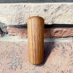 May include: A cylindrical wooden object with a rounded top. The wood grain is visible, and the color is a warm, medium brown. The object is standing upright against a brick wall.