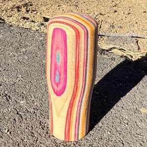 May include: A cylindrical wooden tumbler with a rounded top. The tumbler has a vibrant design with pink, red, orange, yellow, and grey stripes. A pink oval with a turquoise centre is on the front.