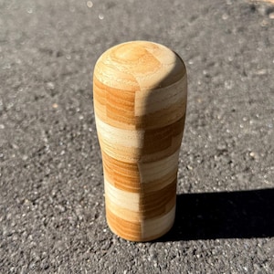 Puede incluir: Un pomo de palanca de cambios de madera con un diseño cilíndrico. El pomo está construido con piezas de madera claras y de tonos medios alternas, creando un patrón de rayas alrededor del cuerpo. La parte superior es redondeada y la base es ligeramente más ancha que el centro.