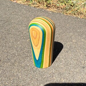 May include: A cylindrical object with a rounded top, crafted from layers of wood in natural tan, bright blue, and vibrant green. The vertical grain pattern is visible throughout the structure, showcasing a smooth, polished finish on the exterior surface.