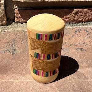 May include: A cylindrical wooden container with a light-colored top and bottom. The body features bands of colorful, striped wood in shades of red, green, blue, and pink. The container is standing on a reddish-brown surface.