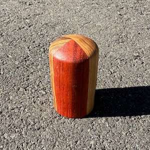 May include: A cylindrical wooden object with a flat top and a rounded base. The object is made of various wood tones, including reddish-brown and light brown. The grain of the wood is visible, and the object appears to be handcrafted.