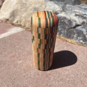 Peut inclure: Un objet en bois cylindrique avec un motif multicolore composé de couches de placage de bois. L'objet est fabriqué à partir de plateaux de skate recyclés.