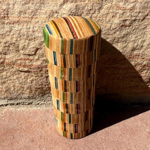 May include: A cylindrical wooden tumbler with a colourful, striped design. The tumbler is made from small, rectangular pieces of wood, with various colours including green, blue, red, and yellow. The tumbler is approximately 15 cm tall.