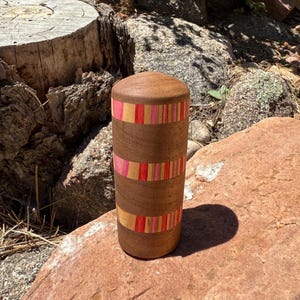 May include: A cylindrical wooden container with a rounded top, featuring a natural brown wood grain finish accented by three horizontal bands of inlaid colorful stripes in shades of pink, red, and light orange, creating a decorative pattern.