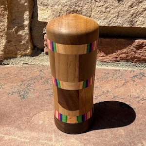 May include: A handcrafted wooden tumbler with a cylindrical shape. The tumbler is made from various wood tones and features colorful bands of rainbow-colored stripes. The top and bottom are a darker wood, and the tumbler is standing on a stone surface.