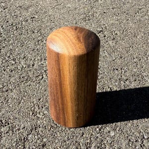 May include: A cylindrical wooden object with a rounded top. The wood grain is visible, showcasing a mix of light and dark brown tones. The object is standing upright on a textured, gray surface, casting a shadow.
