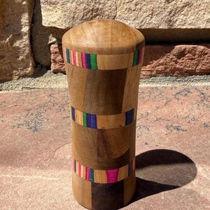 May include: A wooden container with a rounded top. The cylindrical body is made of various wood tones and features three bands of colourful stripes in pink, green, blue, and yellow. The container casts a shadow on the surface.
