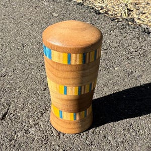 May include: A wooden, cylindrical object with a rounded top and base. The body is light brown with three bands of blue and yellow stripes. The object is standing upright on a textured surface.