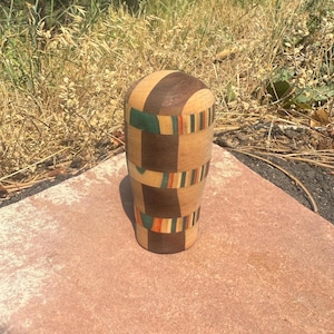 May include: A cylindrical wooden object featuring a segmented design with alternating vertical panels of dark brown, light tan, and thin strips of green, orange, and yellow. The surface is smooth and rounded at the top, showcasing a multi-toned pattern.