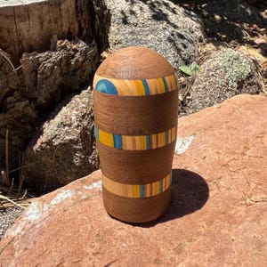 May include: A cylindrical wooden vessel with a rounded top, featuring horizontal bands of natural brown wood grain interspersed with decorative inlay stripes in shades of bright yellow, light blue, and tan, creating a layered pattern.