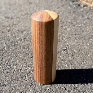 May include: A wooden cylinder with a flat top and a rounded edge. The object is made from two types of wood, with a light-coloured wood on one side and a darker wood on the other. It stands upright on a grey surface.