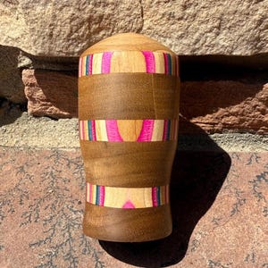 May include: A wooden decorative object with a rounded top and a cylindrical body. The body features bands of light and dark wood, accented with vibrant pink, blue, and yellow stripes. The object is standing on a textured surface.
