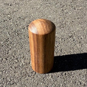 May include: A cylindrical wooden object with a rounded top. The wood grain is visible, showing a warm brown colour. The object is standing upright, casting a shadow on the surface below.