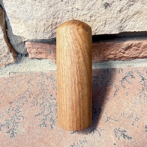 May include: A cylindrical wooden object with a rounded top. The wood grain is visible, and the color is a warm, medium brown. The object is standing upright against a textured stone background.