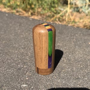 May include: A cylindrical wooden object with a smooth, polished finish. The main body is light brown wood, featuring a vertical inlay strip composed of vibrant purple, bright green, orange, and yellow segments running along the top curve.