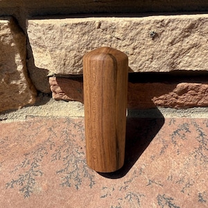May include: A cylindrical wooden object with a rounded top. The wood grain is visible, and the color is a medium brown. The object is standing upright on a reddish-brown surface, with a brick wall in the background.