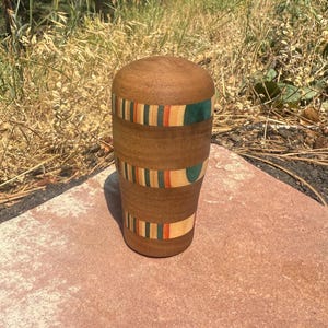 May include: A cylindrical wooden vessel with a rounded top, featuring three horizontal bands of inlaid wood in shades of teal, orange, and light tan, set against the natural brown grain of the main body of the object, which has a smooth finish.