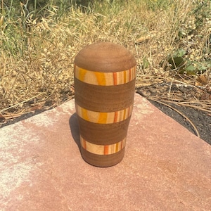 May include: A cylindrical wooden vessel with a rounded top, featuring alternating bands of dark brown wood and lighter wood segments with inlaid stripes of yellow, orange, and red, showcasing a smooth, polished finish and natural wood grain.