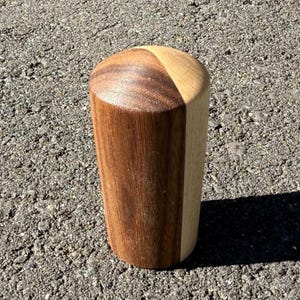 May include: A handcrafted wooden bottle stopper. The stopper is cylindrical with a rounded top. It is made of two types of wood, with a dark brown wood on one side and a lighter wood on the other. The stopper is sitting on a textured surface.