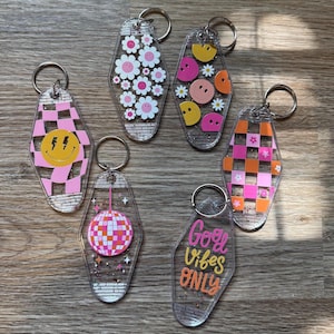 May include: A collection of clear acrylic keychains with various designs. Keychains feature a pink and white checkered pattern with a yellow smiley face, daisies, smiley faces, a disco ball, and the text "Good Vibes Only". Each keychain has a silver keyring.