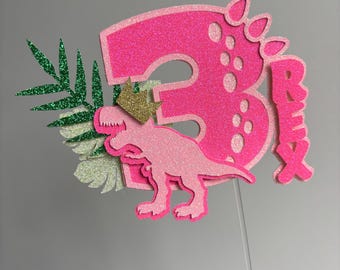 May include: A pink glitter cake topper with the number 3, a dinosaur, and the name "Ruby". The dinosaur wears a gold crown, and green and silver glitter leaves are on the left side. The topper is on a clear stick.