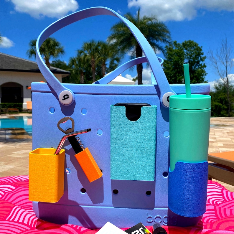 May include: A light blue tote bag with a variety of pockets and accessories. The bag has a built-in bottle opener, a phone holder, and a straw holder. The bag is sitting on a pink and white patterned surface.
