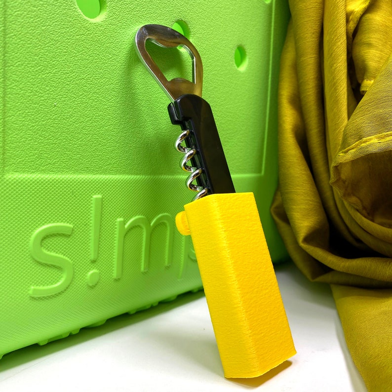 May include: A yellow and black bottle opener with a corkscrew and a bottle opener. The bottle opener is sitting on a green surface with the word "s!m" printed on it.